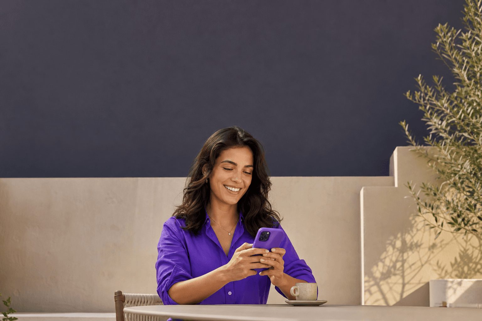 Mulher usando o celular em um café ao ar livre, com camisa roxa, luz natural e arquitetura em tons neutros